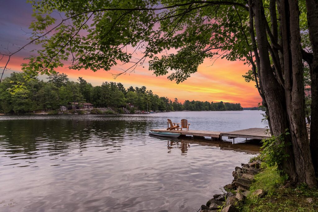 Adirondack chairs on a lake dock at sunset framed by trees and a glowing sky