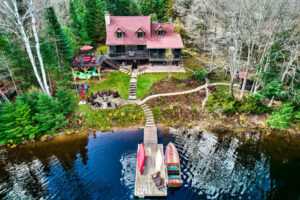 Aerial view of a red-roofed lakefront cottage with dock, fire pit, and forested property