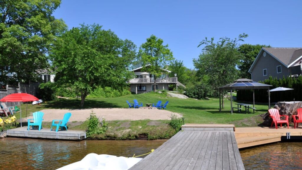 Lakefront cottage with sandy beach, colorful Adirondack chairs, dock, and gazebo on lawn