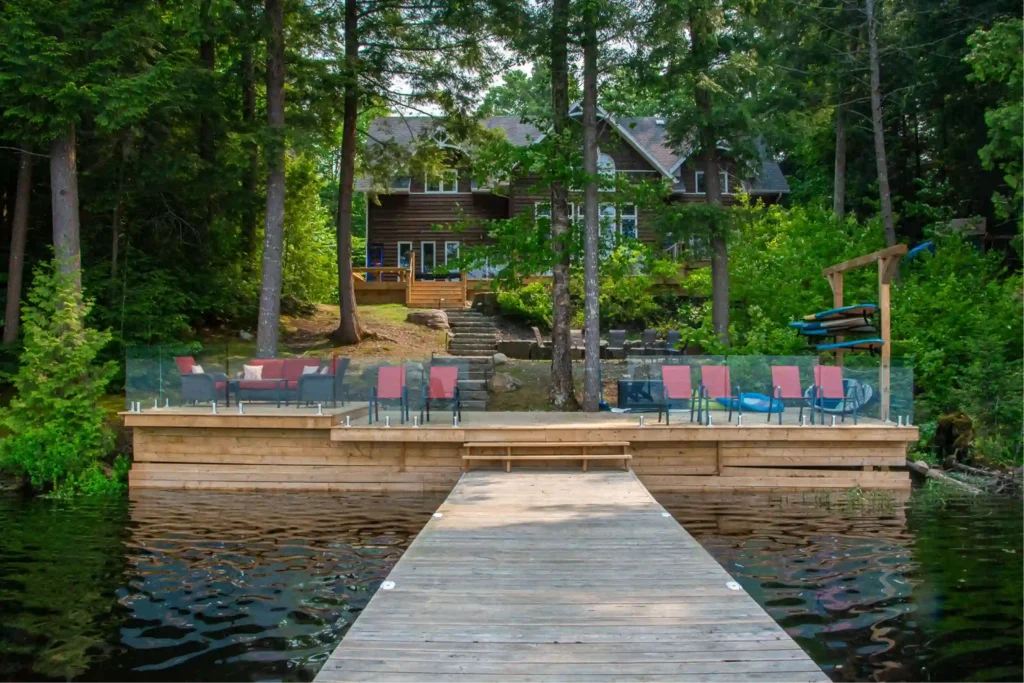 Muskoka cottage rental with private dock, glass-railed waterfront deck, red patio chairs, kayak rack, and stone steps leading to a dark log cottage surrounded by pine trees
