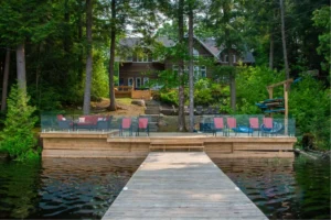 Muskoka cottage rental with private dock, glass-railed waterfront deck, red patio chairs, kayak rack, and stone steps leading to a dark log cottage surrounded by pine trees