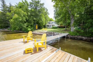 Ontario cottage rental with private dock, bright yellow Muskoka chairs, swim ladder, sandy shallow shoreline, and green lawn leading to a white cottage surrounded by trees