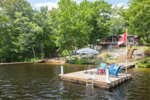 Ontario lakefront cottage rental with private dock, blue Muskoka chairs, patio umbrella, Canadian flag, hammock, children's play set, and screened cottage visible through mature trees