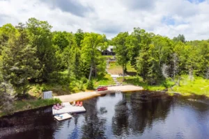 Muskoka cottage rental with private dock, glass-railed waterfront deck, red patio chairs, kayak rack, and stone steps leading to a dark log cottage surrounded by pine trees