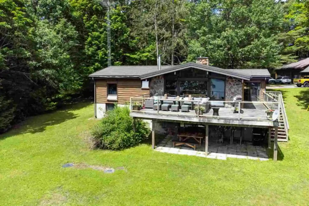Aerial view of an Ontario cottage with stone and wood exterior, large glass windows, furnished upper deck, and covered lower patio on a green lawn