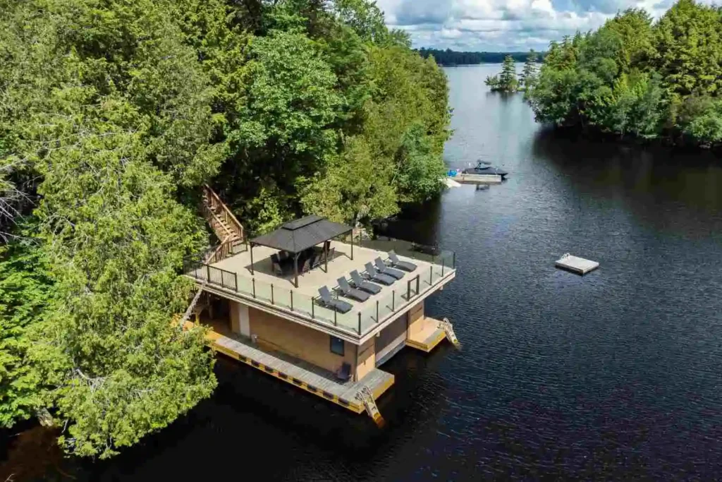Aerial view of a lakefront boathouse with rooftop lounge deck, gazebo, and swim platform on a calm Ontario lake surrounded by forest