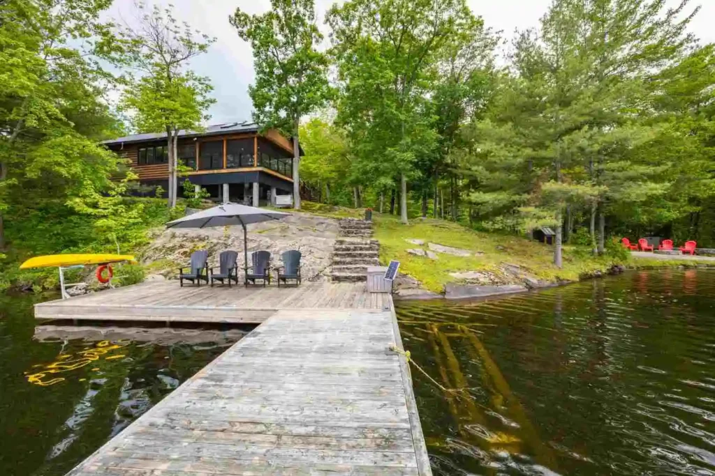 Wooden dock with Adirondack chairs and patio umbrella leading to a modern lakefront cottage surrounded by trees in Ontario
