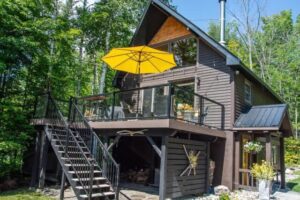 Modern dark chalet with yellow patio umbrella, glass deck railing, and stacked firewood below