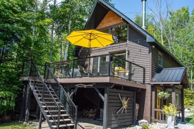 Modern dark chalet with yellow patio umbrella, glass deck railing, and stacked firewood below