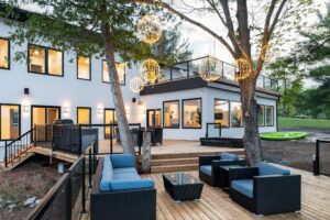 Modern white cottage exterior at dusk with lit deck, outdoor lounge furniture, and globe lights