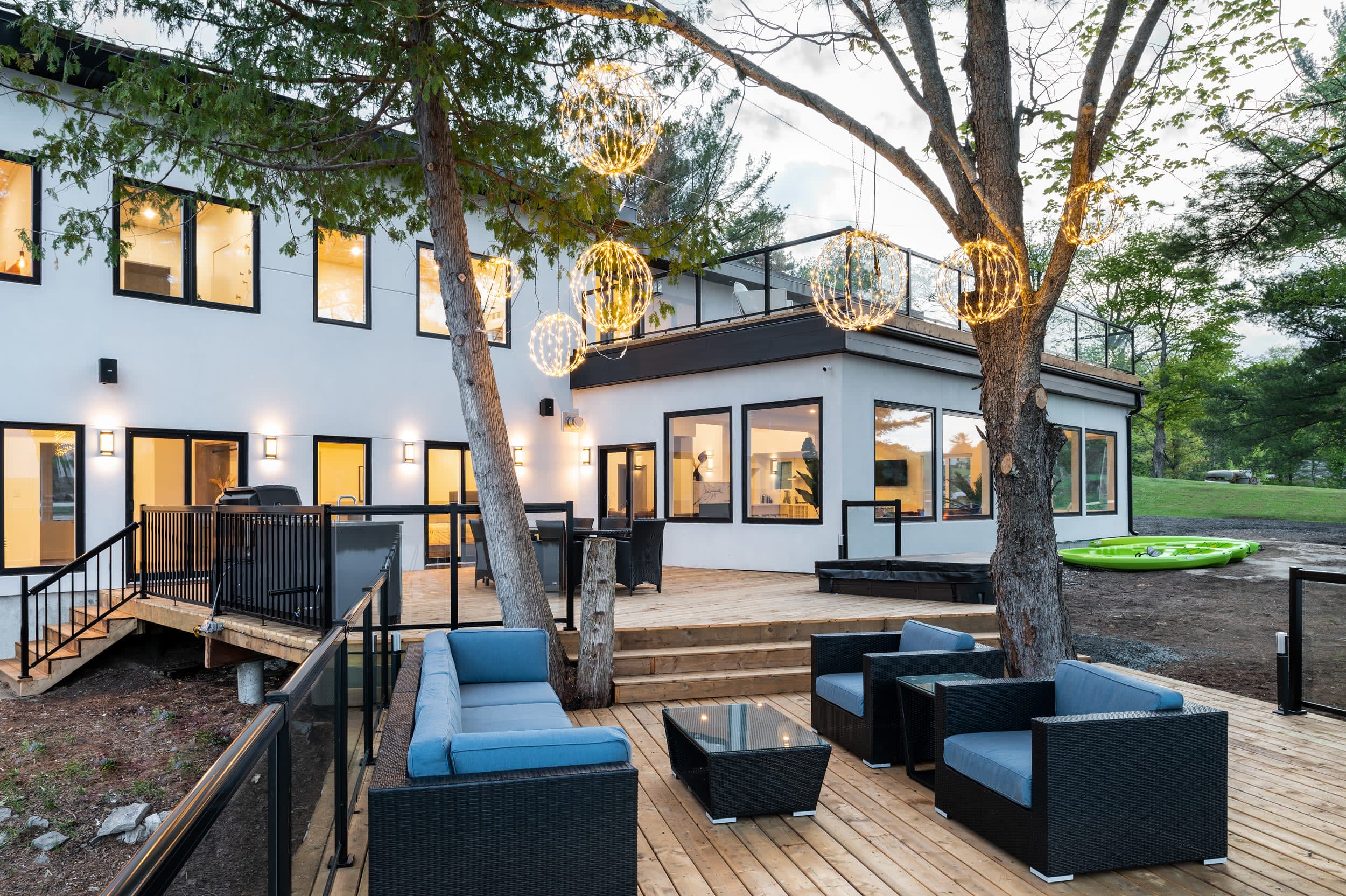 Modern white cottage exterior at dusk with lit deck, outdoor lounge furniture, and globe lights